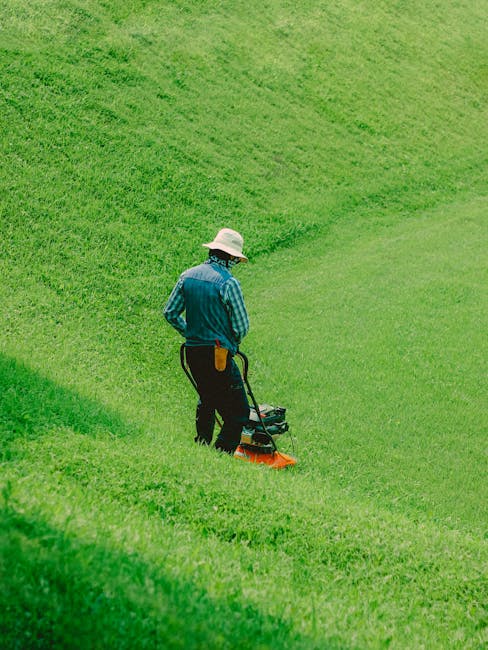 A gardener wearing a wide-brimmed hat, a blue striped shirt, and dark trousers is operating a lawn mower on a gently sloping, well-maintained grassy area. The lush, vibrant green lawn features evenly 